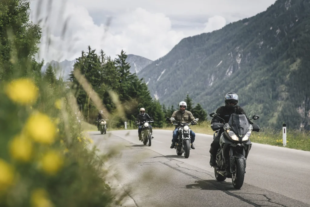 Vier Motorradfahrer auf Alpenstraße mit Bergen und grüner Natur im Hintergrund